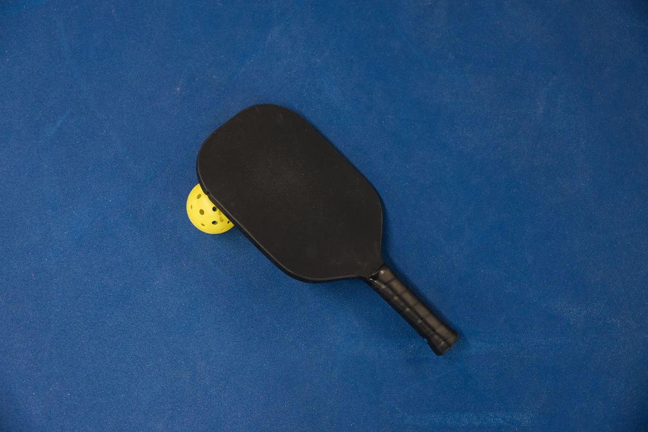 Unbranded black pickleball paddle and yellow ball on a blue court, illustrative stock photograph